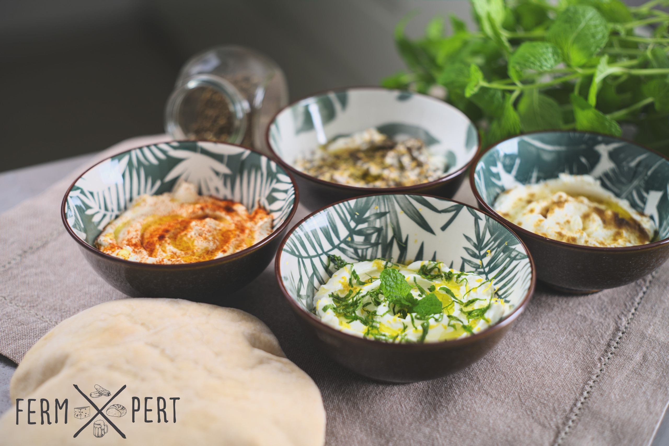 Photo of four bowls of labneh with a pita bread in the foreground and mint leaves in the background