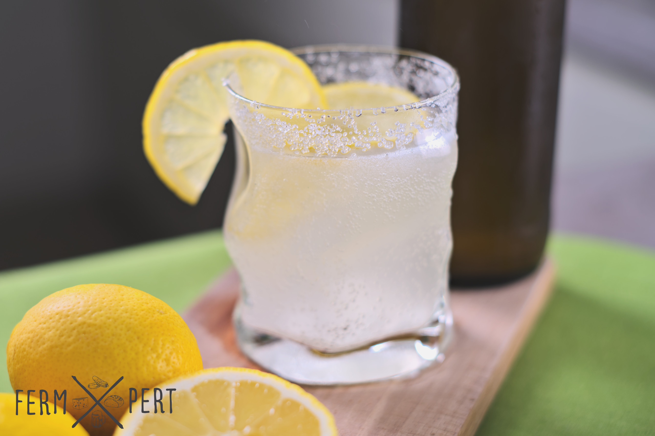 Photo of a glass of lemonade with lemons in front of it and a brown bottle behind.